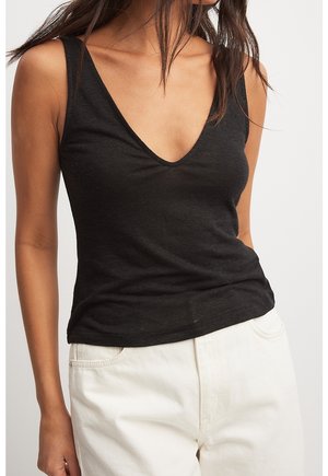 Vrouw draagt een zwarte mouwloze V-hals top en een witte broek, staand tegen een effen lichte achtergrond.
