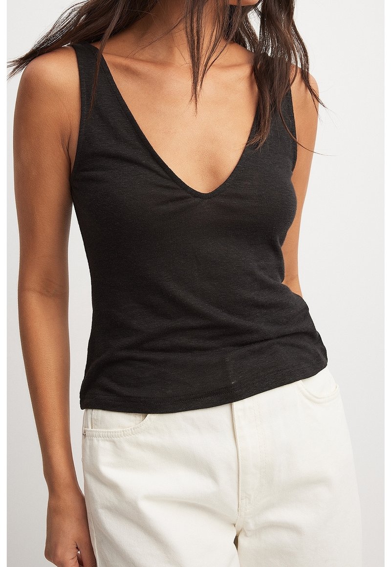Femme portant un haut noir sans manches en V et un pantalon blanc, debout devant un fond clair uni.