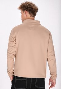 Beige zip-up jakke med en let tekstureret overflade, en lille krave og et overlay-detalje på ryggen, parret med sorte bukser.