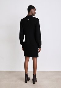 Vestido preto, tricotado, com uma textura canelada, mangas longas e decote redondo. Apresenta um pequeno etiquta na parte de trás e cai até meio do thigh.