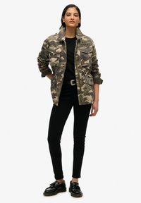 Veste à motif camouflage en olive et marron, dotée de deux poches poitrine et d'un badge de marque, associée à un pantalon noir ajusté et des chaussures noires.