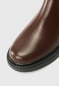 Detailní záběr na hladký hnědý kožený špičku boty s jemným černým prošíváním a silnou černou podrážkou na světlém pozadí.