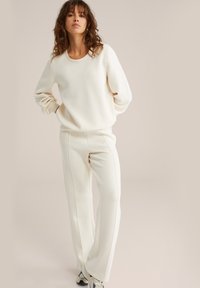 Mujer con cabello rizado, vestida con un suéter crema y pantalones a juego, de pie con las manos detrás de la espalda contra un fondo beige liso.