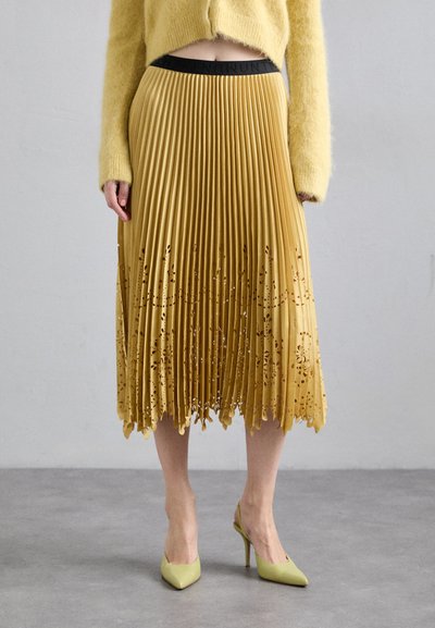 Jupe plissée jaune avec des motifs floraux découpés au laser sur l'ourlet. Dotée d'une taille noire, elle est associée à des escarpins à bout pointu de couleur vert clair.