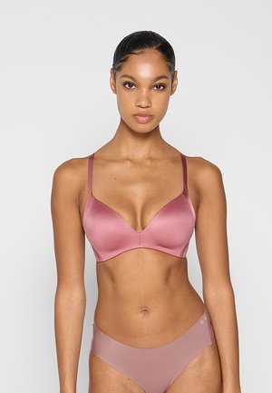 Kobieta z zaczesanymi do tyłu włosami, ubrana w pasujący satynowy biustonosz i majtki w kolorze mauve, stojąca na tle jednolitego jasnoszarego tła.