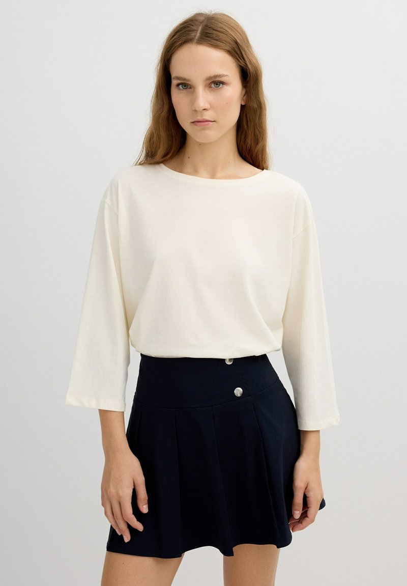 Top de manga larga color crema con escote redondo, combinado con una falda plisada azul marino con acentos de botones plateados. Tejido suave y corte relajado.