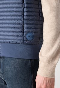 Jūras zila puffer veste ar quiltētu dizainu, elastīgu apakšmalu un Colmar logotipa uzšuvi; apvienota ar bēšu džemperi un tumši ziliem džinsiem.