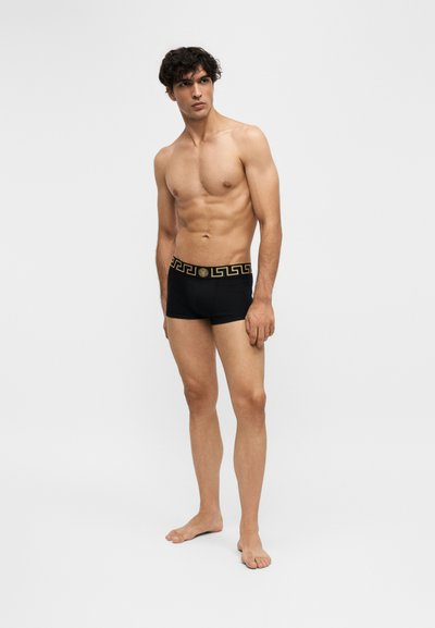 Jeune homme aux cheveux bouclés foncés portant un boxer noir avec une ceinture à motif clé grecque dorée, debout devant un fond clair uni.