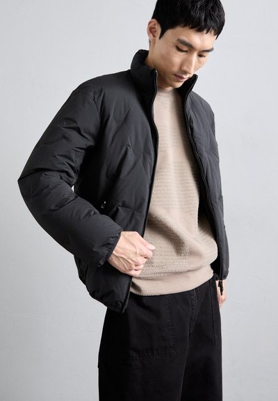 Casaco puffer preto com gola alta, fecho de correr e textura matelassada. Usado sobre um suéter bege com padrão e calças pretas.