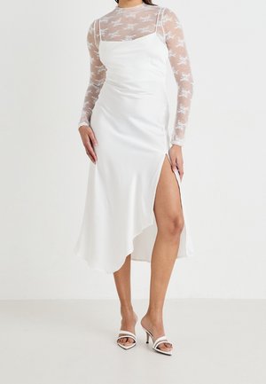 Weißes Satinkleid mit asymmetrischem Saum und einem seitlichen Schlitz. Kombiniert mit einem transparenten Langarmshirt mit floralem Spitzenmuster.