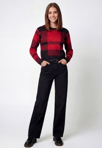 Red and black checkered knitted sweater with a rounded neck, paired with black high-waisted pants and black shoes, featuring a belt.