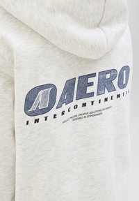 Haut à capuche gris clair, tissu doux, avec un imprimé graphique bleu au dos portant la mention "AERO INTERCONTINENTAL" et un détail de logo.