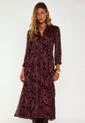 Femme portant une robe midi à manches longues, motif cachemire bordeaux, avec des détails froncés et des bracelets dorés au poignet.