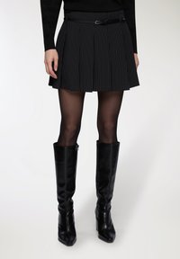 Donna che indossa una mini gonna plissettata nera a righe, collant trasparenti neri e stivali alti neri in pelle su uno sfondo bianco.