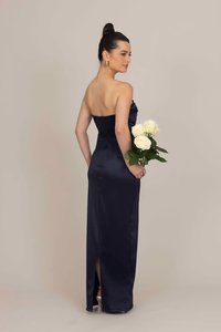 Αμάνικη ναυτική σατέν τουαλέτα με εφαρμοστή σιλουέτα, διαθέτοντας σχισμή και πλαϊνές ρυτίδες. Το μοντέλο κρατά λευκά τριαντάφυλλα με το ένα χέρι.