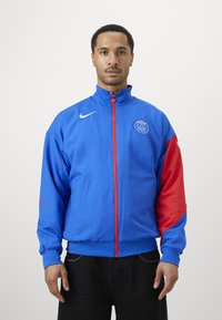 Blue and red tracksuit jacket made from lightweight fabric, featuring a full zipper, elastic cuffs, and a logo on the chest and sleeve.
