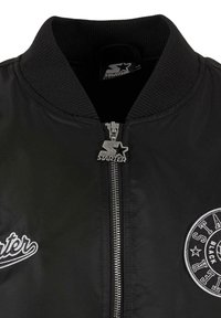 Veste bomber noire dotée d'un col côtelé, d'une fermeture éclair argentée et d'un logo brodé sur le côté gauche de la poitrine. Tissu lisse et léger.