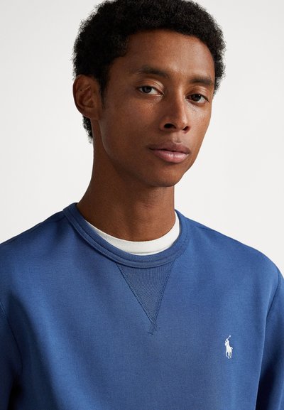 Zils kokvilnas džemperis ar apaļu kakla izgriezumu, uz krūtīm baltā izšūta logo un ap kaklu teksturēts apdare.