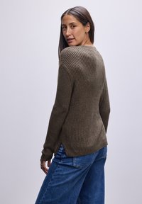 Olivgrüner gerippter Pullover mit langen Ärmeln, der einen Schlitzsaum und einen taillierten Körper aufweist. Kombiniert mit blauen weit geschnittenen Jeans.