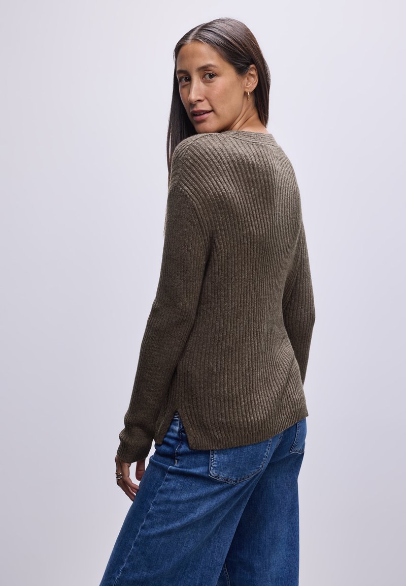 Olivgrüner gerippter Pullover mit langen Ärmeln, der einen Schlitzsaum und einen taillierten Körper aufweist. Kombiniert mit blauen weit geschnittenen Jeans.