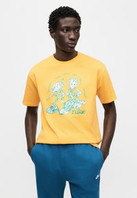 Πορτοκαλί βαμβακερό μπλουζάκι με πράσινο γραφικό σχέδιο που παρουσιάζει χαρακτήρες και κείμενο. Στρογγυλή λαιμόκοψη και κοντά μανίκια, συνδυασμένο με μπλε φόρμες.