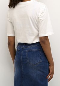 Camiseta de algodón blanca de manga corta, vista desde la parte posterior, combinada con una falda de mezclilla azul que presenta bolsillos traseros y un corte recto.