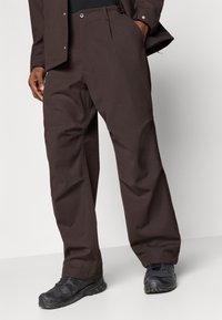 Dark brown trousers made of textured fabric, featuring a relaxed fit, front pleats, and a button closure. Paired with black shoes.
