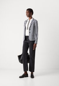Blazer con motivo houndstooth blu e bianco, abbinato a jeans neri a gamba larga e mocassini neri. La modella tiene una borsa nera.