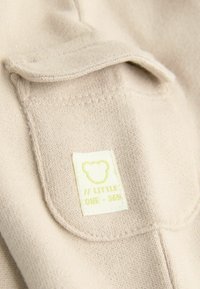 Beige kötött anyag texturált felülettel, elülső zsebével, valamint fehér címk�ével, amelyen sárga medve logó és a "LITTLE ONE - 365" felirat található.