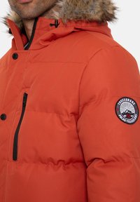 Homme portant une veste d'hiver orange avec une capuche bordée de fourrure, une poche poitrine zippée et un écusson rond sur la manche indiquant « Threadbare Polar Technology ».