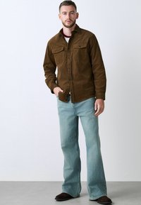 Brown corduroy shirt jacket with two chest pockets, worn over a white and maroon shirt, paired with light blue flared jeans and dark shoes.