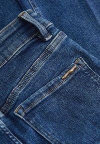 Blaue Jeans aus Denim mit einer strukturierten Oberfläche, ausgestattet mit orangefarbenen Nähten, einem Gürtelschlaufen und einem kleinen Metallelement an der Tasche.