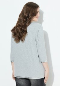 Frau mit langen, welligen braunen Haaren, die ein hellgraues 3/4-Ärmel-Oberteil und dunkle Hosen trägt. Sie steht vor einer weißen Wand und ist leicht zur Seite gedreht.
