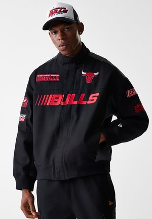 Zwarte jas met rode "BULLS" letters en geborduurde logo's, met een hoge kraag en geribde manchetten, gecombineerd met een wit en zwart pet.