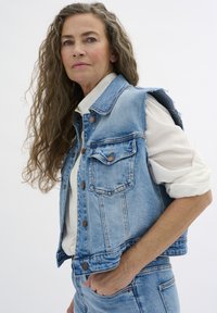 Modrý denimový vest s roztrhnutými ramenami, prednými vreckami a hnedými gombíkmi. Nosený cez bielu blúzku, podčiarkujúci neformálny vrstvený vzhľad.