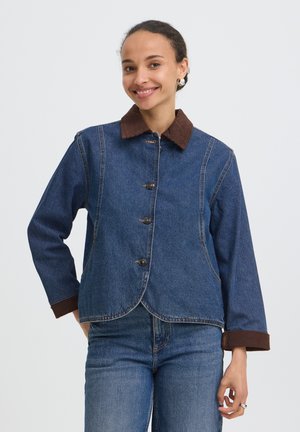 Femme souriante portant une veste en denim bleue avec un col et des poignets marron, associée à un jean bleu, debout devant un fond uni.