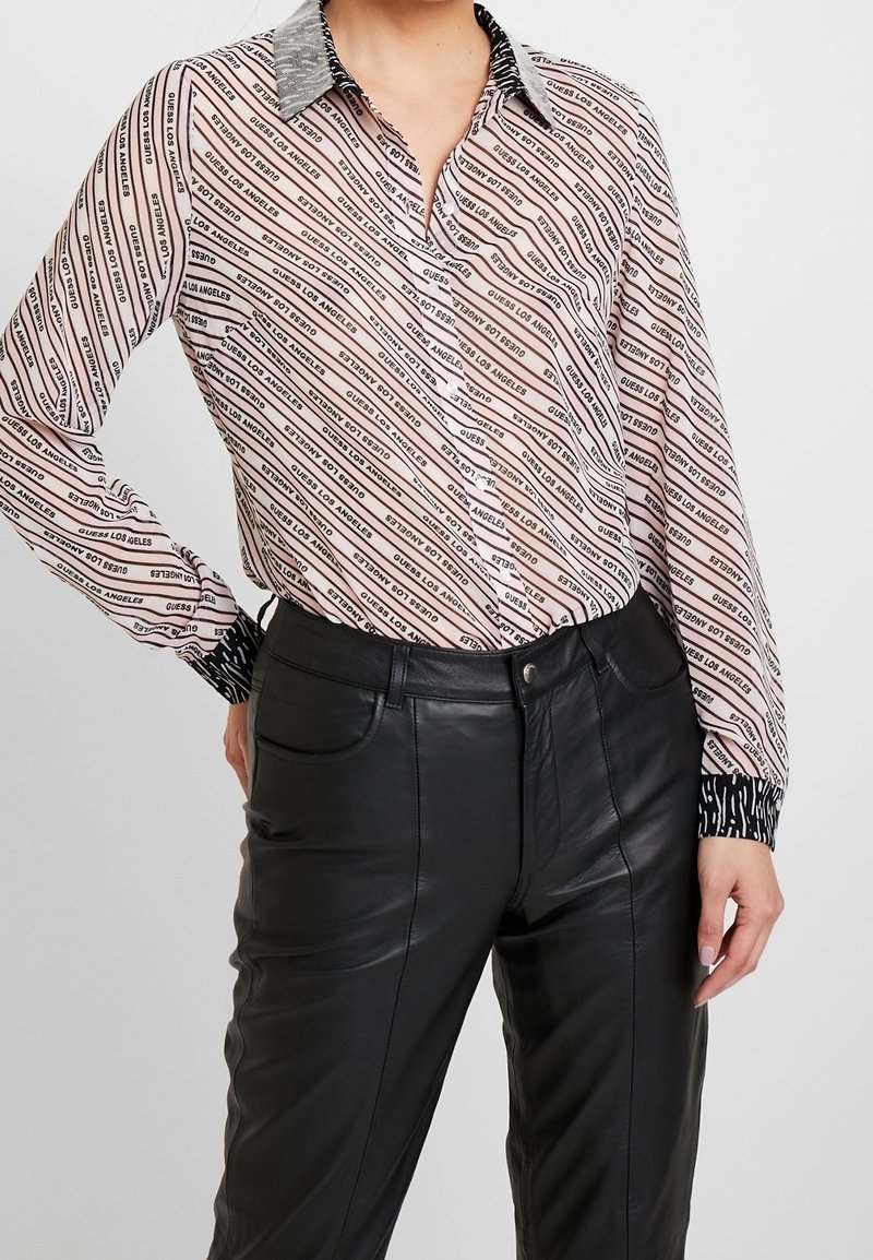 Personne portant une chemise rose à rayures avec le texte "Guess Los Angeles" et un pantalon noir en simili cuir, debout devant un fond uni.