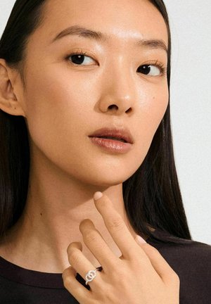 Silver ring with interlocking design, worn on a finger. The model has smooth skin and dark hair, wearing a dark top.