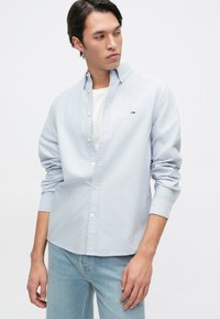 Jasnoniebieska koszula z długim rękawem, z zapięciem na guziki, okrągłym kołnierzykiem i haftowanym logo. Nosi się ją na białym T-shircie, w zestawie ze jasnymi dżinsami.