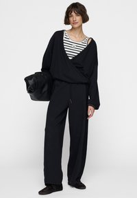 Zwarte oversized sweatshirt met brede mouwen, gecombineerd met zwarte sweatpants en een gestreept tanktop. Het model houdt een zwarte leren tas vast.