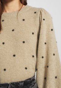 Pull beige en tissu doux avec des pois noirs, doté d'un col rond côtelé et de manches bouffantes.