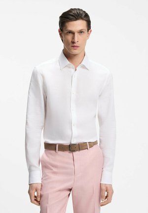 Mann trägt ein tailliertes weißes Hemd mit Knöpfen, das in hoch taillierte rosa Hosen mit braunem Gürtel gesteckt ist, vor einem schlichten Hintergrund.