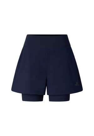 Ung kvinde iført hvid kasket, hvid 'FIRE NICE' t-shirt, marineblå shorts og chunky hvide sneakers, poserer mod en ensfarvet baggrund.