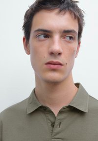 Olijfgroene polo met een knoopsluiting bij de kraag, gemaakt van zachte katoen stof; bevat een subtiele naaddetail vlakbij de halslijn.