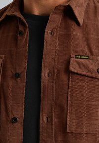 Bruine corduroy shirt met een ruitpatroon, voorzien van zwarte knopen, een zak met een label, en gedragen over een zwart shirt. Structuurafwerkings.