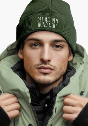 Green knit beanie with a stitched patch reading "DER MIT DEM HUND GEHT." The wearer is in a light green puffer jacket with a black interior.