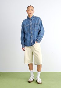 Jeansknappskjorte med to brystlommer, beige knelengde shorts, hvite sokker og brune sneakers mot en lys bakgrunn.