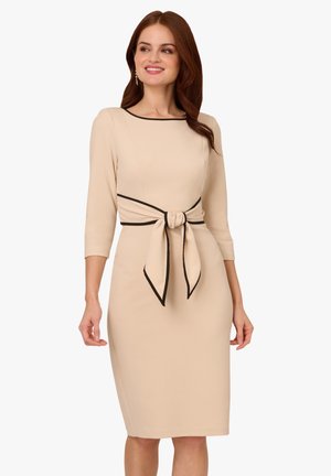 Béžové šaty se třičtvrtečními rukávy, přiléhavým střihem a černou stuhou v pase. Hladká látka s kontrastními okraji.