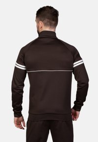 Veste zippée marron foncé avec des rayures blanches d'accentuation sur les épaules. Présente un col haut et des poignets et ourlet côtelés. Texture lisse.