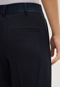 Close-up van een persoon in zwarte, op maat gemaakte broek met een verborgen achterzak en een elastische tailleband met het merk "JOOP!"